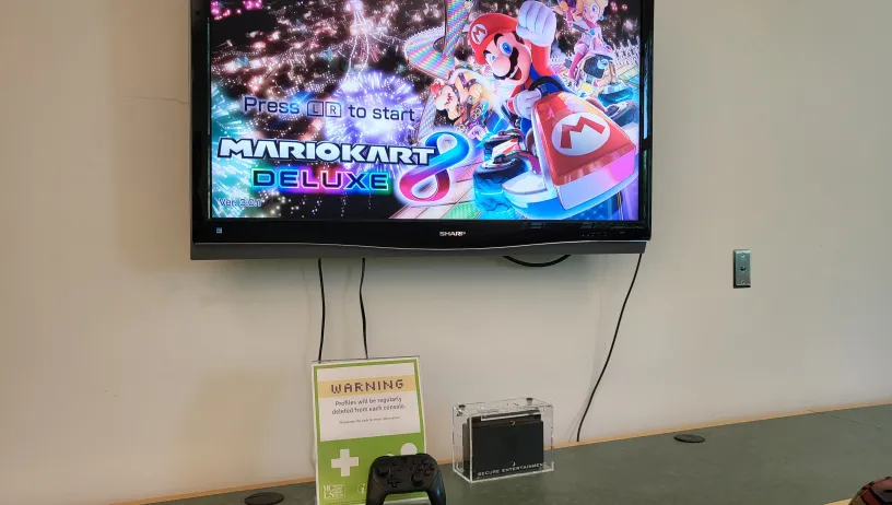 The image shows a television screen mounted on a wall, displaying the title screen of the game "Mario Kart 8 Deluxe." Below the screen, on a green counter, there is a clear plastic box containing a Nintendo Switch console. Next to the box, there is a black game controller. A sign with a warning about profile deletion from the console is also present on the counter. The area appears to be set up for gaming, likely in a public or communal space. 