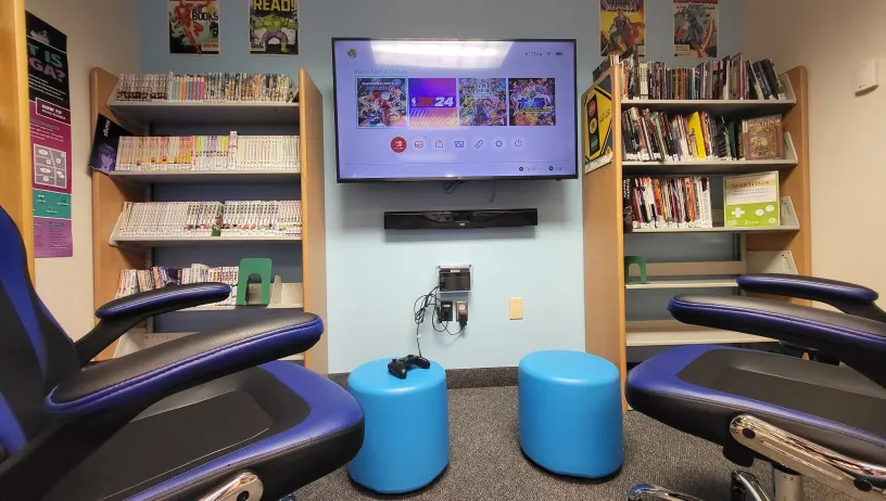  This image depicts a cozy and well-equipped gaming space at the Hoke Library, featuring comfortable seating with blue and black chairs, and an interactive digital display showing a Nintendo Switch game menu. The walls are adorned with shelves filled with a diverse collection of graphic novels, books, and video games. The presence of the Nintendo Switch enhances the room's appeal, making it a vibrant and inviting environment for visitors to enjoy both gaming and reading. 