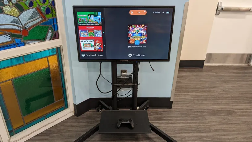 The image shows a television screen mounted on a stand with a Nintendo Switch connected to it. The television screen displays a user interface of a gaming console, likely a Nintendo Switch.On the right side of the screen, there is an option to "Continue" with a game that appears to be "Super Smash Bros. Ultimate."  Below the television, there is a shelf attached to the stand, which holds a gaming controller. The stand is placed against a wall, and to the left of the stand, there is a colorful stained-glass.
