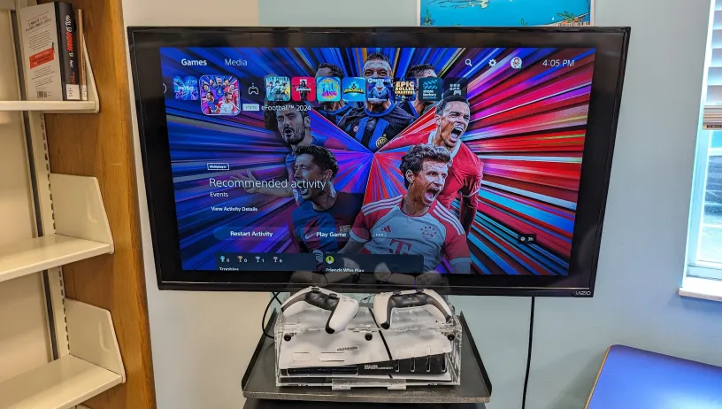 The photo shows a PlayStation 5 gaming setup at the Elisabeth Lahti Library. A flat-screen TV is mounted on a wall or stand, displaying the PlayStation 5 home screen. The screen features the game "eFootball 2024" with various other game icons visible in the top menu, such as "Fall Guys," "Stray," and "Epic Roller Coasters." The time on the screen reads 4:05 PM.  Below the TV, there is a clear acrylic stand holding a PlayStation 5 console and two DualSense controllers. The controllers are white and black.