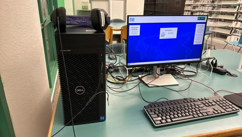 A Dell Precision 3660 desktop computer setup with a monitor, keyboard, mouse, and headphones on a desk in a library. The screen shows a blue background with text.