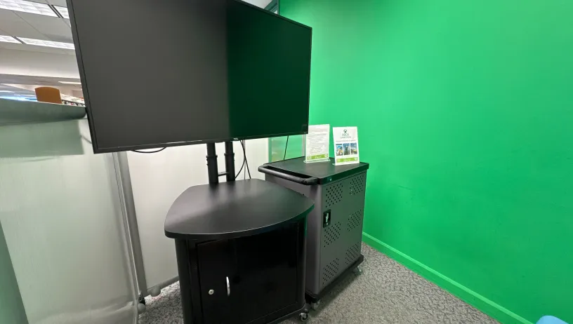 The image shows a corner setup for gaming or presentations. A large flat-screen TV is mounted on a stand, with a black cabinet underneath that has a small door. Next to the TV stand, there is a black equipment cart with a sign on top, likely providing instructions or information about the setup. The wall behind the cart is painted bright green, resembling a green screen backdrop. The area is well-lit, and part of an office or communal space is visible in the background.