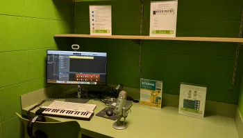 The image shows a small studio with green walls, equipped with a computer, MIDI keyboard, Yeti microphone, and headphones on a desk.