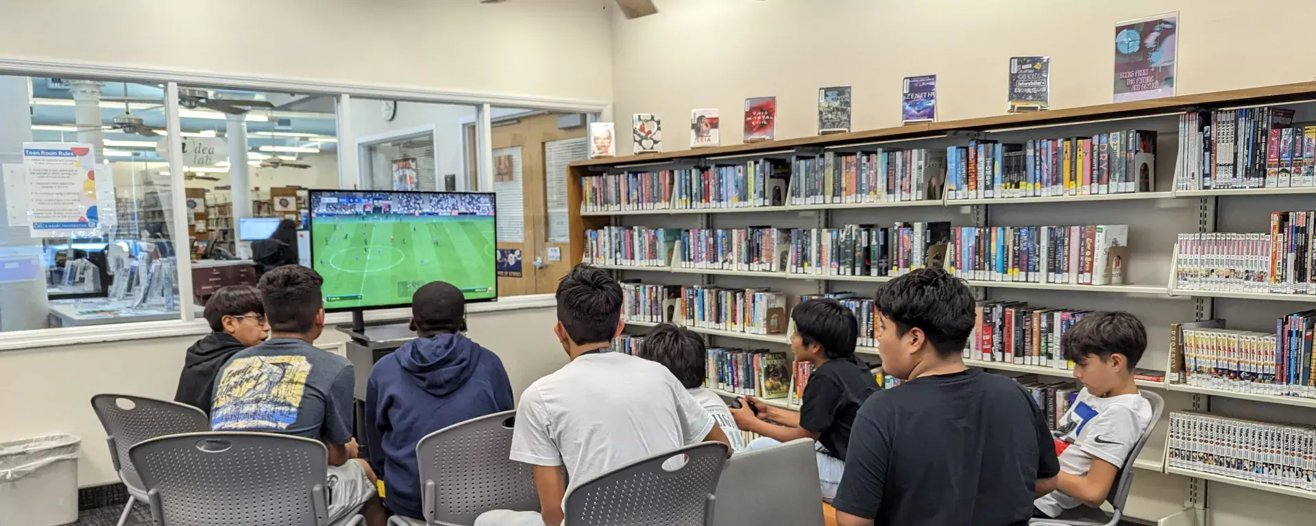 a group of young people in a library, situated in a room filled with bookshelves that are stocked with a wide variety of books. They are seated on metal chairs, arranged in a semi-circle facing a television screen displaying a soccer game. The atmosphere seems casual and comfortable, indicating a community space that encourages leisure activities alongside reading. There are books prominently displayed on the top of the shelves, and the environment suggests a well-maintained public space.