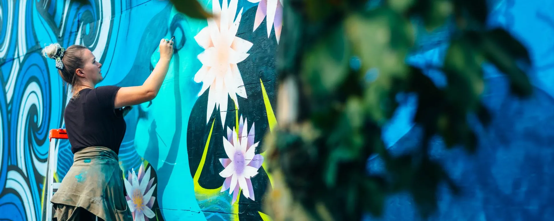 Image of a woman painting a mural of water lilies on an outdoor wall of a building.