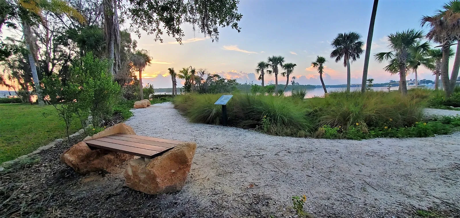 Image of site four of the Ripple Project that faces the river and features lush greenery and a walking path with benches during sunrise.